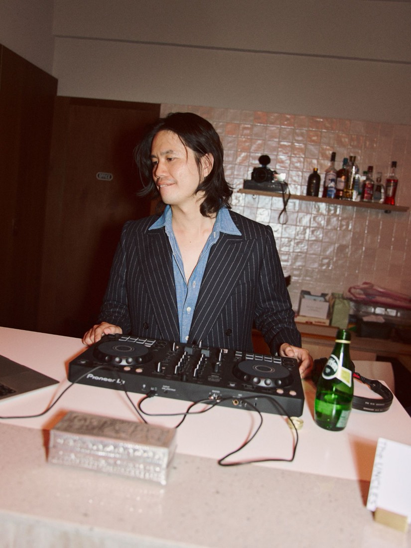 Gene 穿著細條紋西裝站在 DJ 台後，身旁是 Perrier 氣泡水
