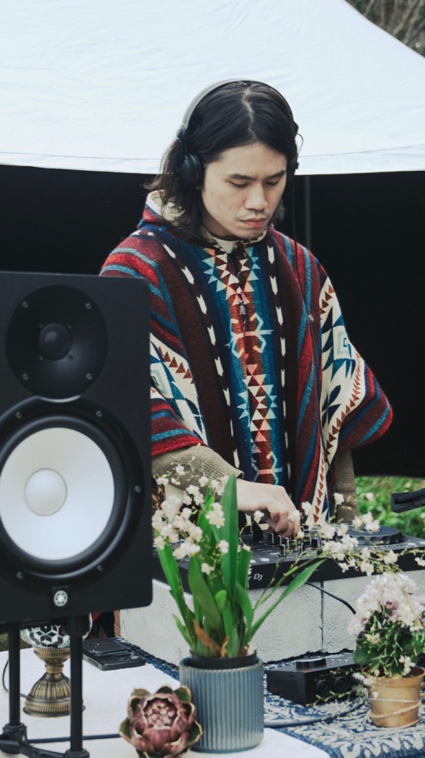 Gene 穿著披風、戴耳機專注混音，前方有花草與監聽喇叭